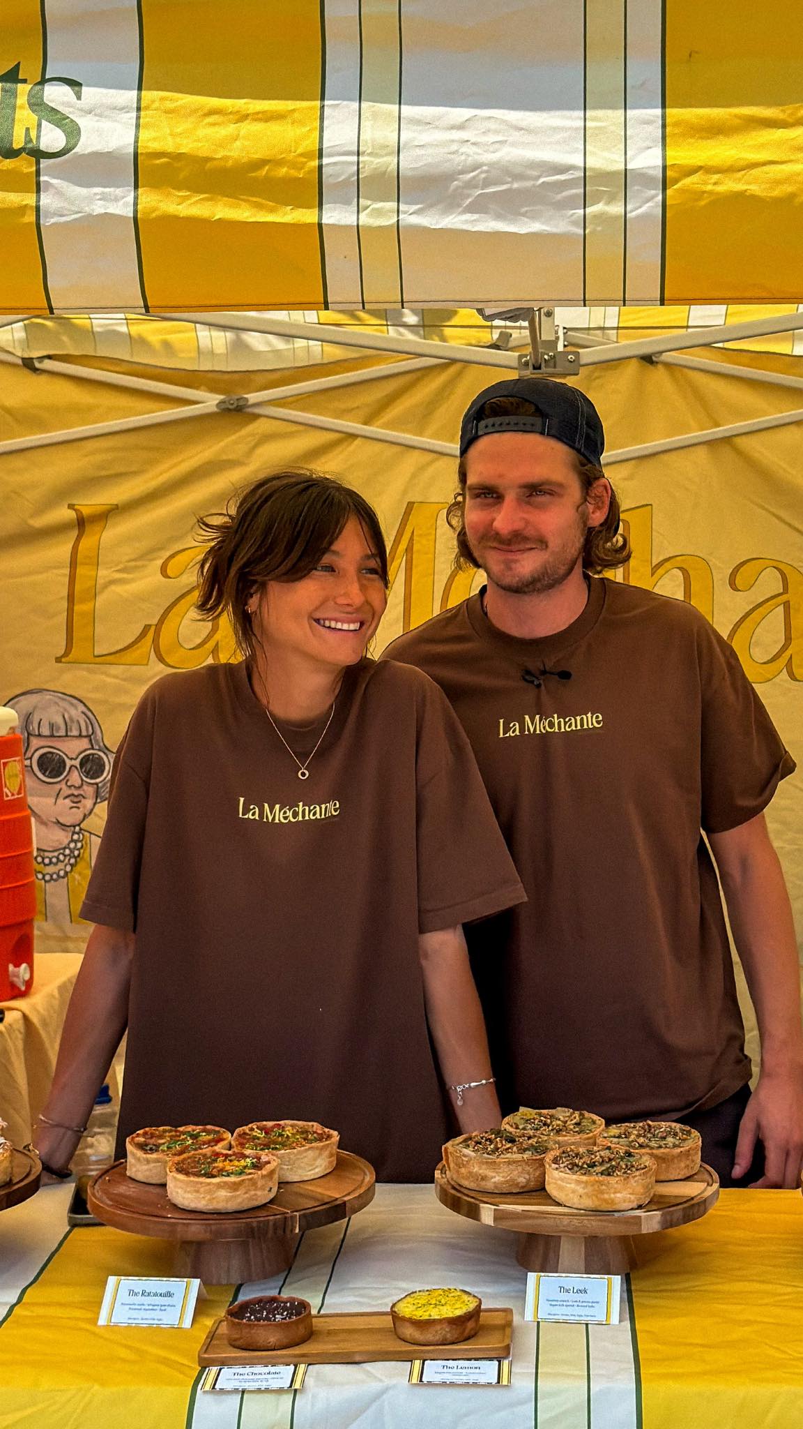 Julie and Samuel in La Mechante aprons behind a market stall of savoury tarts, under a yellow-striped awning.