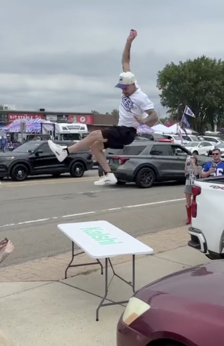 A tailgater mid-air above a Kalshi-branded folding table in a parking lot, fist raised, with the crowd watching.