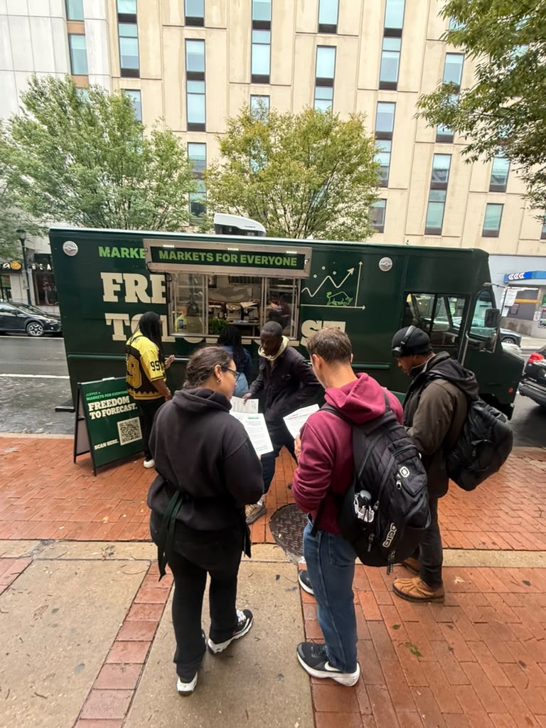 A green Freedom to Forecast coffee truck reading Markets for Everyone, parked on a city sidewalk, with people reading flyers beside a sign with a QR code.