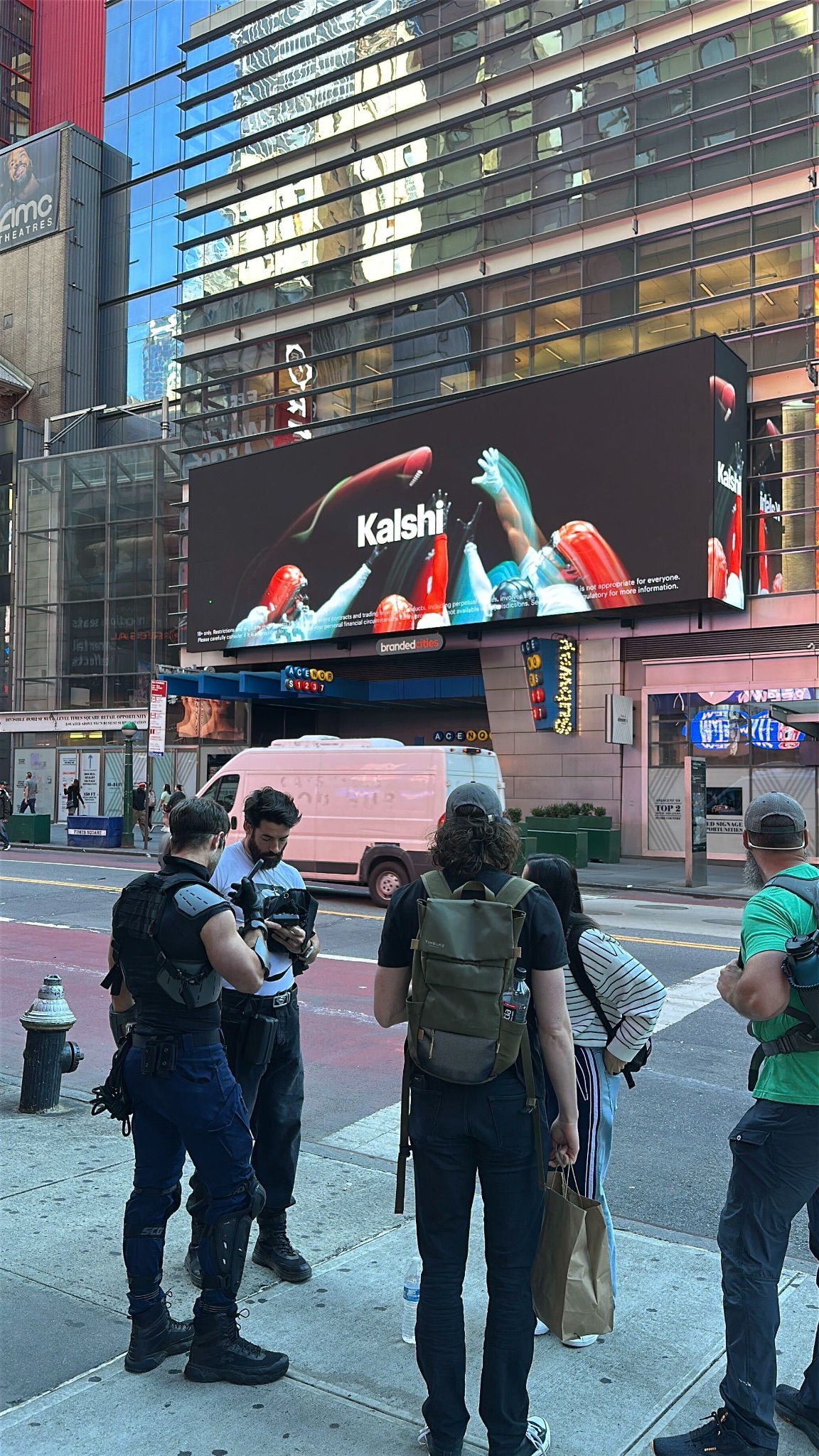 A Kalshi billboard in Times Square showing football players leaping for a ball, above a street corner where people stand checking their phones.