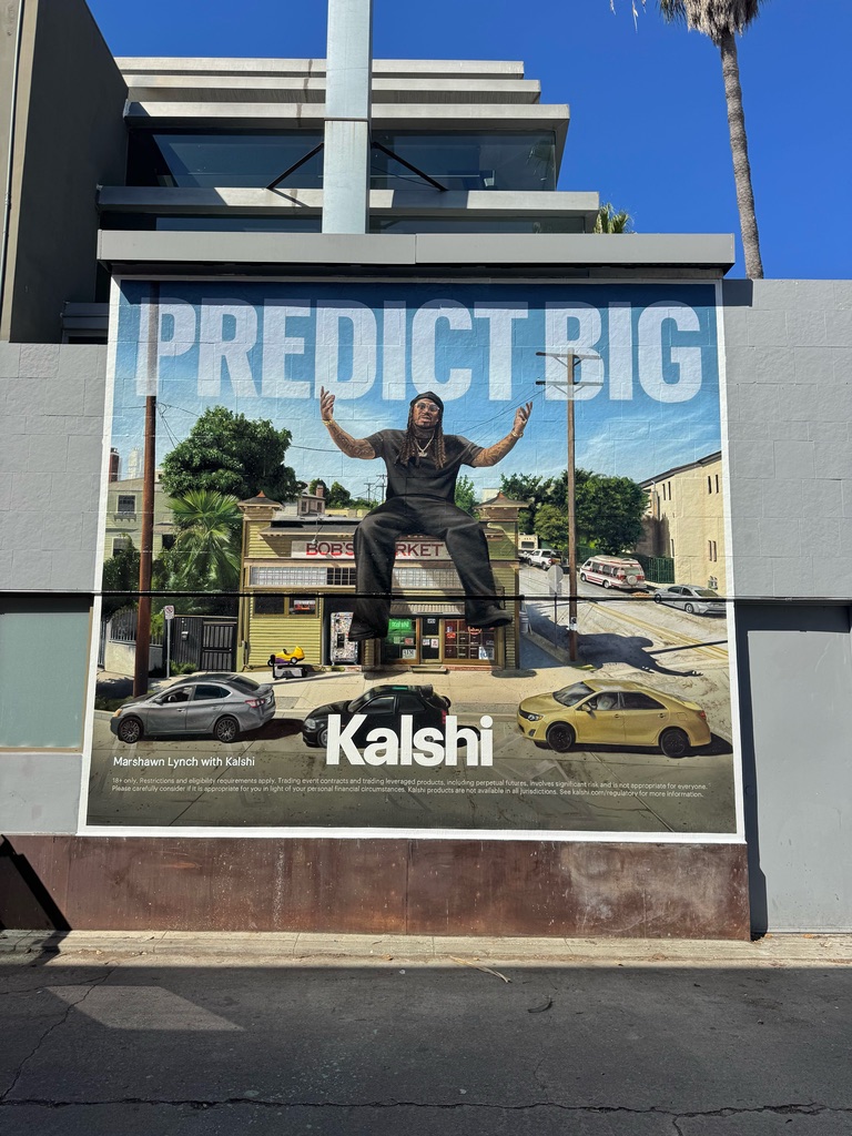 A Kalshi mural reading Predict Big, with Marshawn Lynch sitting on top of a corner market, painted on a wall under a palm tree.